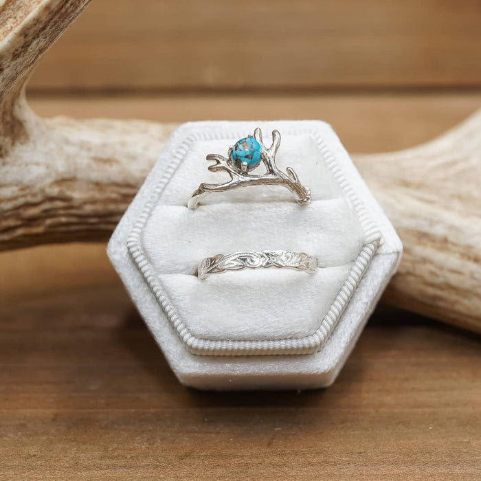Two silver rings are displayed on the 2 Slot Velvet Ring Box - Promo*, which features a striking hexagonal shape. The top ring is adorned with a blue gemstone and an antler-inspired design, while the bottom ring boasts an intricately engraved pattern. Antler decor accents rest nearby on a rustic wooden surface, creating an enchanting scene ideal for wedding photography or proposal photo shoots.