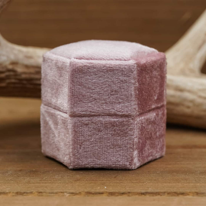The Velvet Hexagon Ring Box, featuring a soft pink velvet exterior, rests on a wooden surface with an antler in the background, creating an elegant scene ideal for wedding photography or proposal photo shoots.