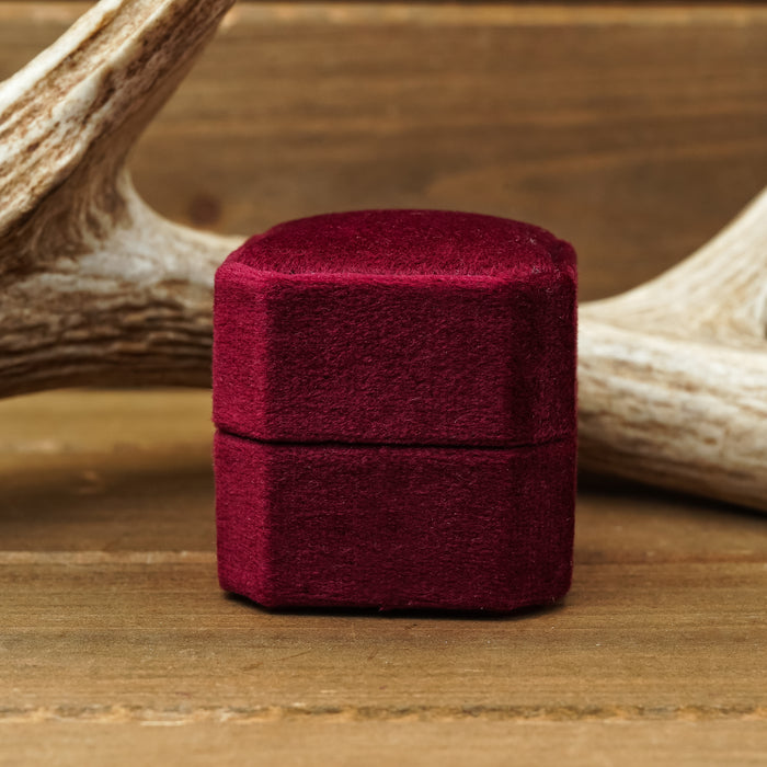 A compact, closed 2 Slot Velvet Ring Box - Promo* in rich burgundy velvet rests on a wooden surface with a deer antler in the background—an elegant detail often featured in wedding photography and proposal photo shoots.