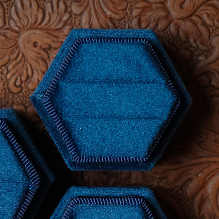 A 2 Slot Velvet Ring Box in hexagonal blue velvet, ideal for elegant wedding photography, sits on a brown embossed surface with a floral pattern.