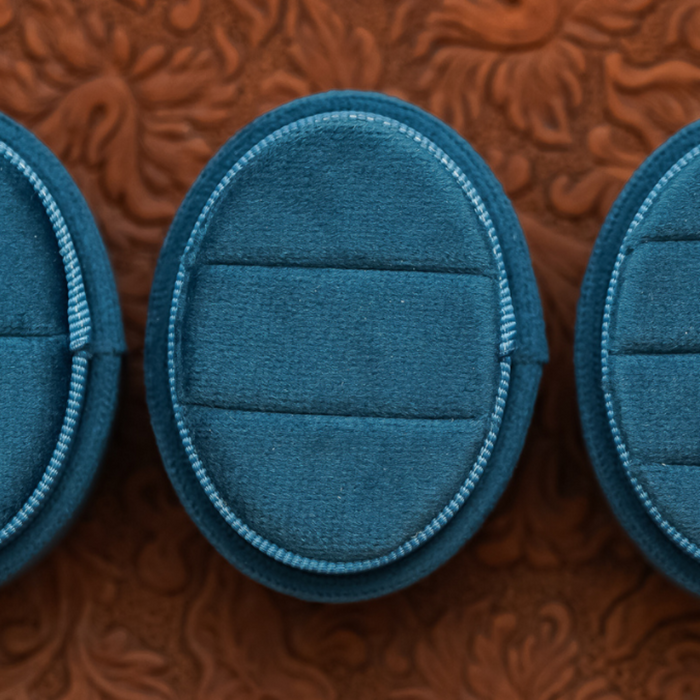 A close-up of the 2 Slot Velvet Ring Box - Promo* in blue velvet, featuring an oval shape and two ring slots, is displayed closed on an embossed brown surface. Ideal for wedding photography or proposal photo shoots, the empty box sits with two similar velvet ring boxes partially visible on either side.