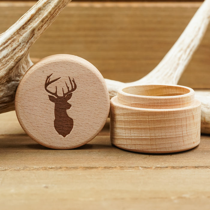The Antler Ring Gift Box is a small beech wood ring box featuring a lid engraved with a deer head silhouette, shown resting on a wooden surface beside antlers.