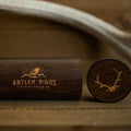 A dark wooden ring box with Antler Rings and an antler logo engraved on the side, paired with a matching lid featuring an antler illustration, rests on a wooden surface beside a stylish Antler Sunglasses Case for your favorite antler sunglasses.