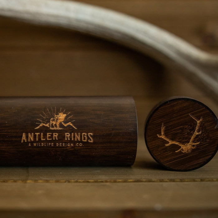 A dark wooden ring box with Antler Rings and an antler logo engraved on the side, paired with a matching lid featuring an antler illustration, rests on a wooden surface beside a stylish Antler Sunglasses Case for your favorite antler sunglasses.