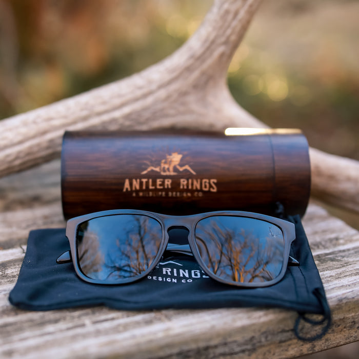 Antler Sunglasses are shown reflecting trees and sky, resting on a branded pouch in front of the Antler Sunglasses Case—a stylish bamboo accessory—set against antlers with a softly blurred outdoor background.