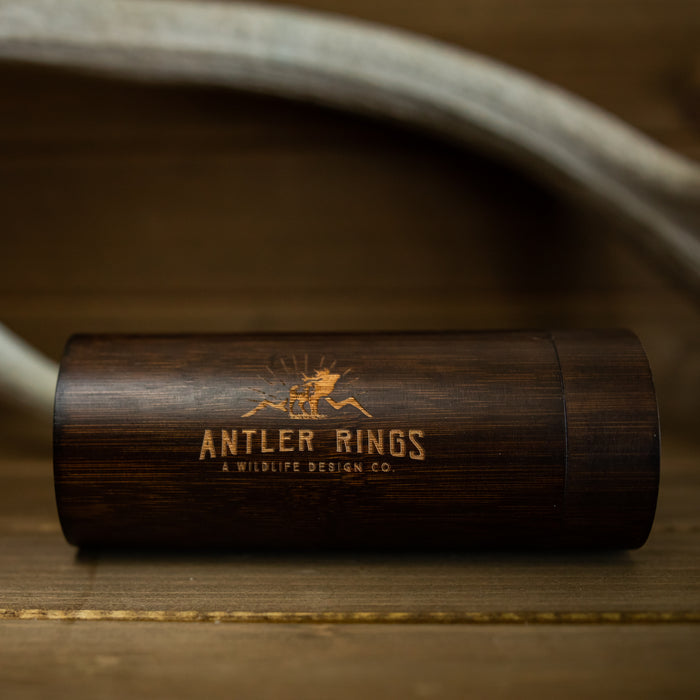 A cylindrical wooden Antler Sunglasses Case by Antler Rings: A Wildlife Design Co., featuring an engraved antler logo, rests on a wooden surface with a white antler in the background—perfect for storing your favorite antler sunglasses or eyewear.