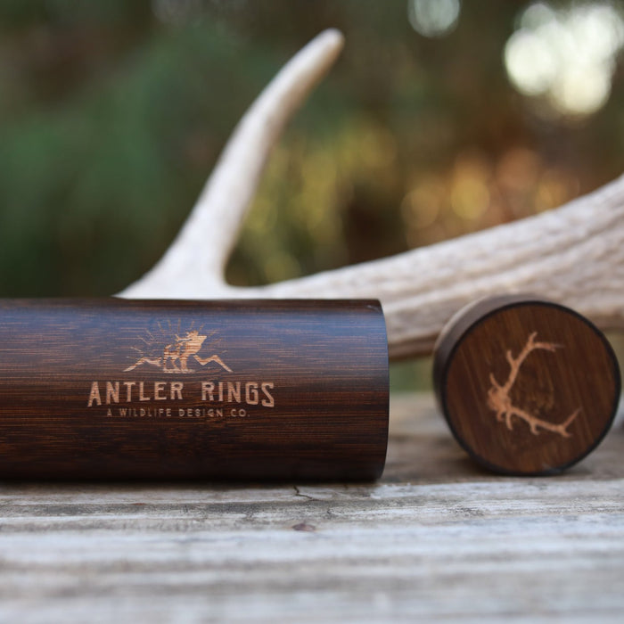 A wooden ring box with Antler Rings and an antler logo engraved on it sits on a rustic wooden surface beside an antler and the Antler Sunglasses Case, with blurred greenery in the background.