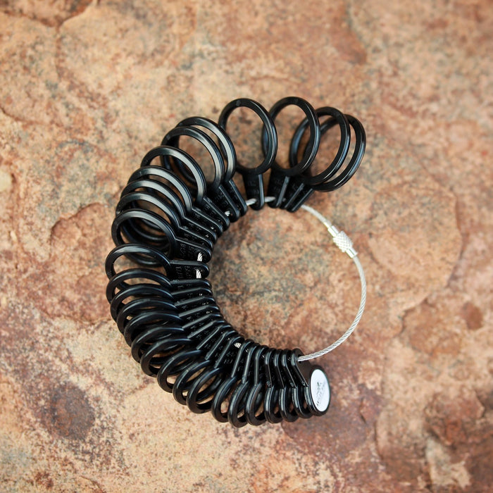 The AntlerRings Ring Sizer features a metal ring holding black plastic sizing bands arranged in a semicircle, forming a practical tool for precise ring size measurement, displayed on a textured brown stone surface.