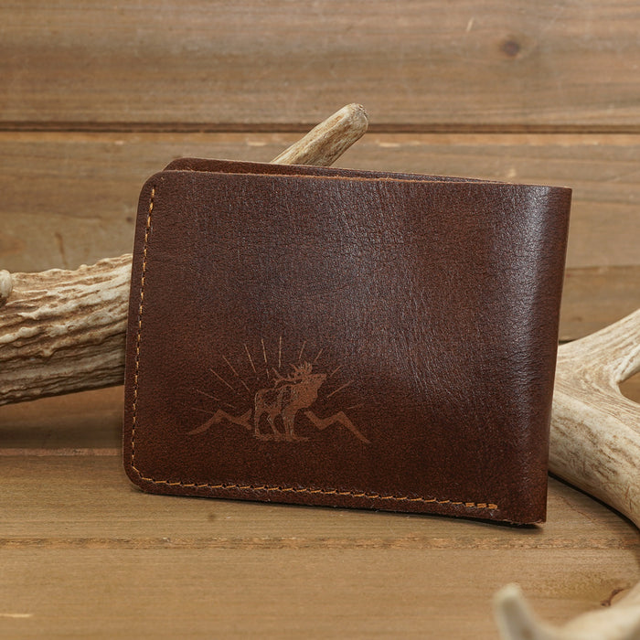 A Deer Leather Wallet featuring a moose and mountain design rests on a wooden surface, propped against antlers—a perfect outdoorsman accessory that blends rustic charm with practicality.