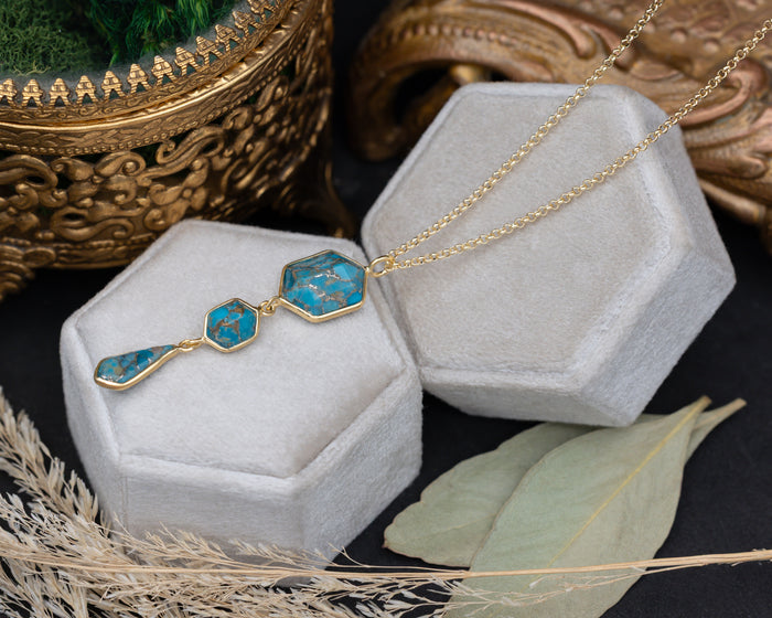 A gold pendant necklace with three turquoise stones on a gold chain, sitting on a white cushion with dried flowers in the background