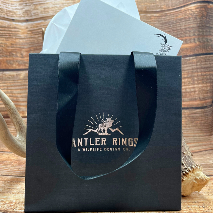 A "Gift Packaging" bag with black ribbon handles, labeled “Antler Rings: A Wildlife Design Co.,” rests on a wooden surface. Antlers and white tissue paper appear in the background, creating an elegant scene of thoughtful gift presentation.