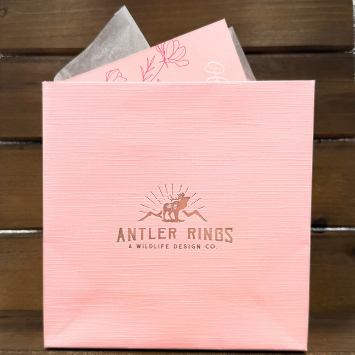 Gift Packaging: A pink gift bag featuring the Antler Rings: A Wildlife Design Co. logo, elegantly presented with tissue paper and a card decorated with floral designs, all set against a wooden background.