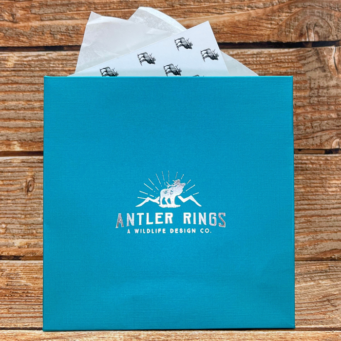 A blue Gift Packaging box with white tissue paper sticking out, placed on a wooden surface. The box features a stag logo and the text Antler Rings: A Wildlife Design Co., lending an elegant touch to your gift presentation.