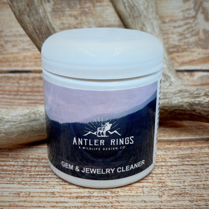 A white container labeled Jewelry Cleaner rests on a wooden surface with antlers in the background. Ideal for fine jewelry, this non-toxic cleaner features a label decorated with a mountain and antler design.