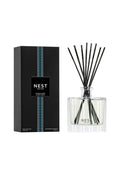 A reed diffuser bottle with black reeds next to its black packaging box, both featuring the NEST New York logo and a blue vertical stripe design, on a clean white background.