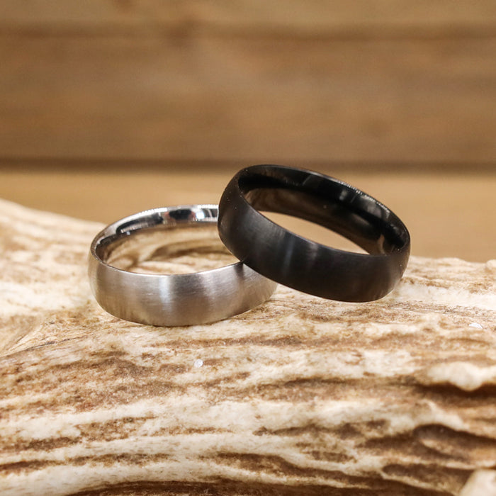 Two Stainless Steel Wedding Bands rest on a piece of textured wood—one featuring a polished silver finish with brushed detailing, and the other in a matte black design. The blurred wooden background highlights the Activity Bands in the foreground.