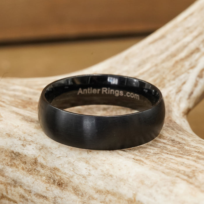 A Stainless Steel Wedding Band - Activity Band with a men’s matte black brushed finish rests on a light-colored antler. The inside of the ring is engraved with the text AntlerRings.com, while the background appears blurred and brown.