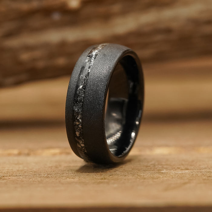 The Allende, a matte black tungsten ring featuring a textured, rough central inlay, stands upright on a wooden surface with a blurred wooden background.