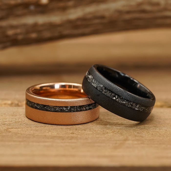 Two rings from “The Allende” collection rest on a wooden surface: one is copper-colored, the other is matte black tungsten. Both feature a central inlay of rough, dark stone evocative of meteorite. The background shows blurred wood grain.