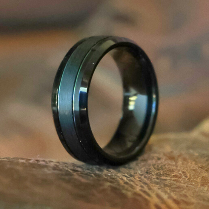 The Bandera, a black tungsten ring featuring a subtle central groove, stands upright on a textured brown surface. Its sophisticated brushed finish highlights the enduring appeal of this men's tungsten ring against a softly blurred background.