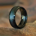 The Bandera, a black tungsten ring featuring a subtle central groove, stands upright on a textured brown surface. Its sophisticated brushed finish highlights the enduring appeal of this men's tungsten ring against a softly blurred background.