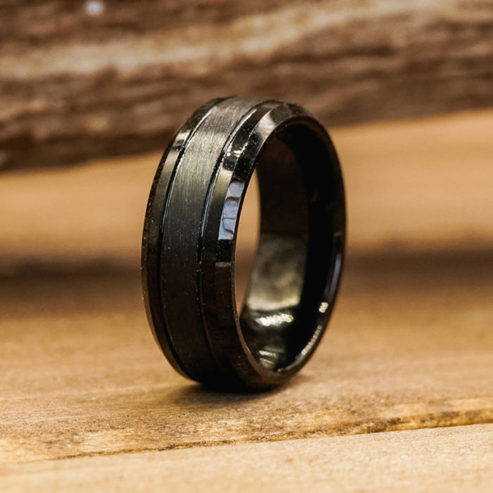 The Bandera, a men's black tungsten ring featuring a brushed center and polished beveled edges, stands upright on a wooden surface, showcasing its modern elegance.