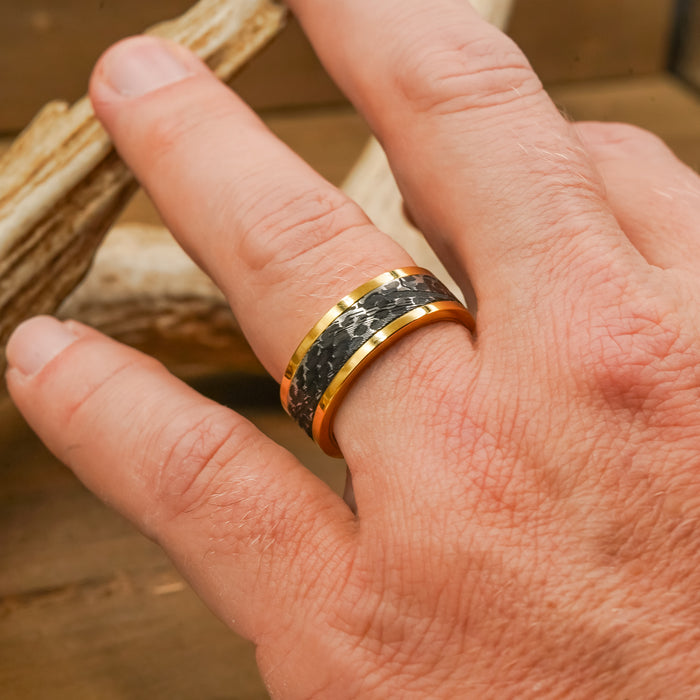 A hand wearing The Bannack gold men’s ring, featuring a striking black pattern, rests on a wooden surface with part of an antler visible in the background.