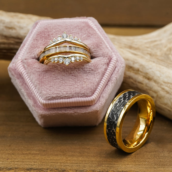 Three gold rings with diamonds are displayed in a pink velvet hexagon box, their brilliance enhanced by the soft blush tones. The Bannack—a thick gold men’s ring featuring a dark, hammered brushed band—rests on the wooden surface nearby, adding a modern touch. In the background, a piece of antler completes the elegant arrangement.