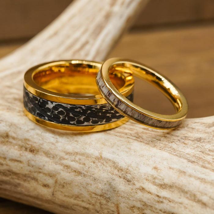 Two gold rings rest on a deer antler. One is The Bannack, a striking gold men’s ring featuring a wide black patterned Damascus steel inlay, while the other showcases a thin, light gray textured inlay reminiscent of a hammered brushed ring. Both are set against a blurred wooden background.