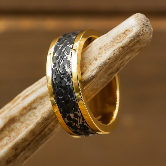 The Bannack, a gold men’s ring featuring a textured black inlay inspired by a hammered brushed design, is displayed on light-colored wood set against a brown background.