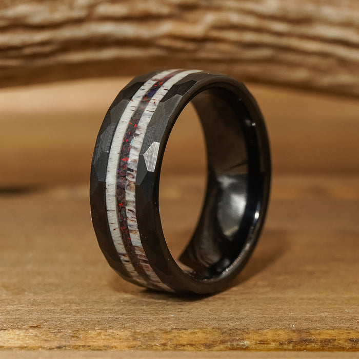The Benton is a black tungsten carbide ring with a faceted surface, featuring two inlaid stripes—one white and the other red-speckled—displayed against a rustic wooden background.