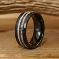 The Benton is a black tungsten carbide ring with a faceted surface, featuring two inlaid stripes—one white and the other red-speckled—displayed against a rustic wooden background.