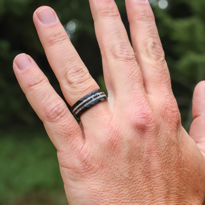 A hand wearing The Benton, a bold black tungsten carbide ring accented with silver stripes, is shown on the ring finger against a blurred green outdoor background.