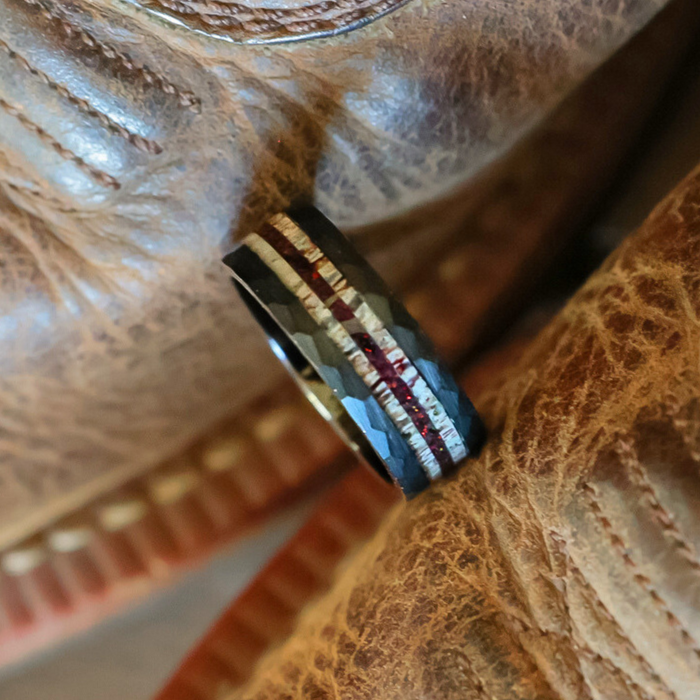The Benton, a black tungsten carbide ring featuring a faceted texture and two inlaid stripes—one silver and one red—rests on worn brown leather.