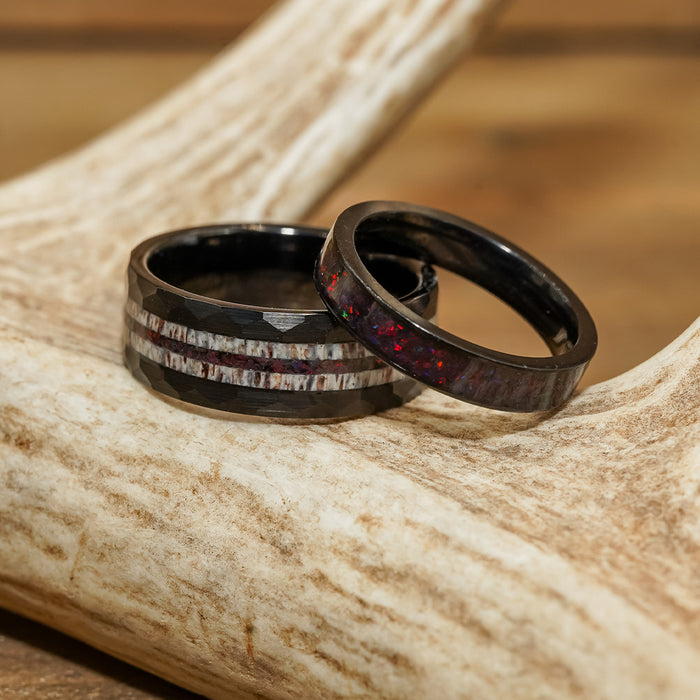 Two black rings with inlaid designs—one is The Benton, a striking black tungsten carbide ring featuring a strip of iridescent red, while the other is an antler inlay ring with a lighter, textured band—rest gracefully on a light-colored, textured antler.