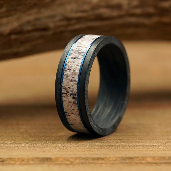 A close-up of The Berkshires men's wedding band, showcasing a black carbon fiber ring with a textured light-colored inlay and thin blue accent lines, standing upright on a wooden surface against a blurred wood background.