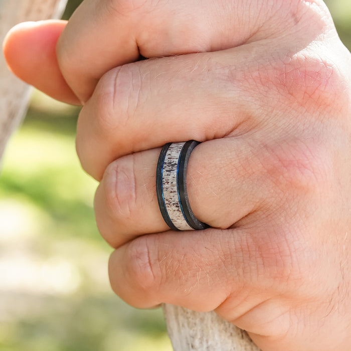 A close-up of a hand gripping a wooden object, wearing The Berkshires men's wedding band, which features a dark edge and a speckled, textured center reminiscent of an antler ring. The background is blurred greenery.