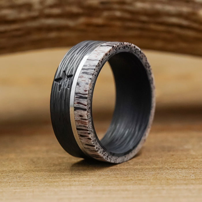 A close-up of The Blackwater Falls men's 9mm ring, highlighting its textured design with a dark inner band, silvery metal stripe, and light, wood-like outer edge—displayed on a wooden surface against a softly blurred wooden background.