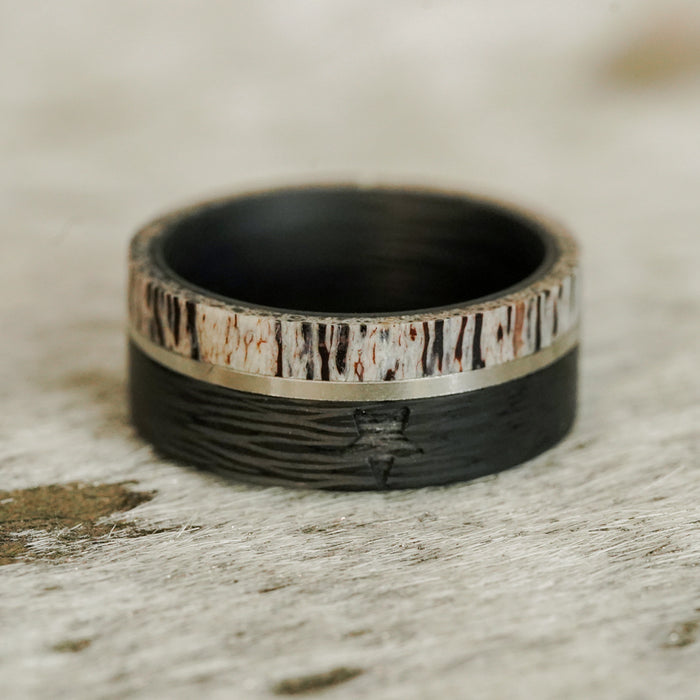 A close-up of The Blackwater Falls men's 9mm ring, showcasing its textured black base and a top band crafted from genuine antler with natural brown and white patterns. The antler ring rests on a light, textured surface.