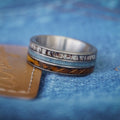A close-up of The Bodacious—a multi-layered men’s band crafted from tungsten carbide, featuring silver, blue, brown, and patterned inlays with antler ring accents—resting on denim fabric beside a tan leather patch.