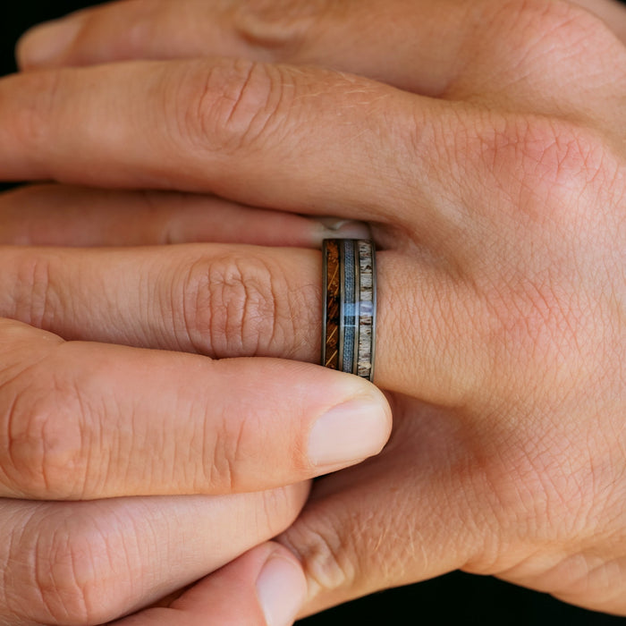 A close-up of hands, with one wearing The Bodacious—a distinctive men's band featuring multiple inlaid bands of different colors and textures. This ring showcases unique elements such as wood, blue, and stone-like segments, reminiscent of finely crafted antler rings and enhanced by the durability of tungsten carbide.