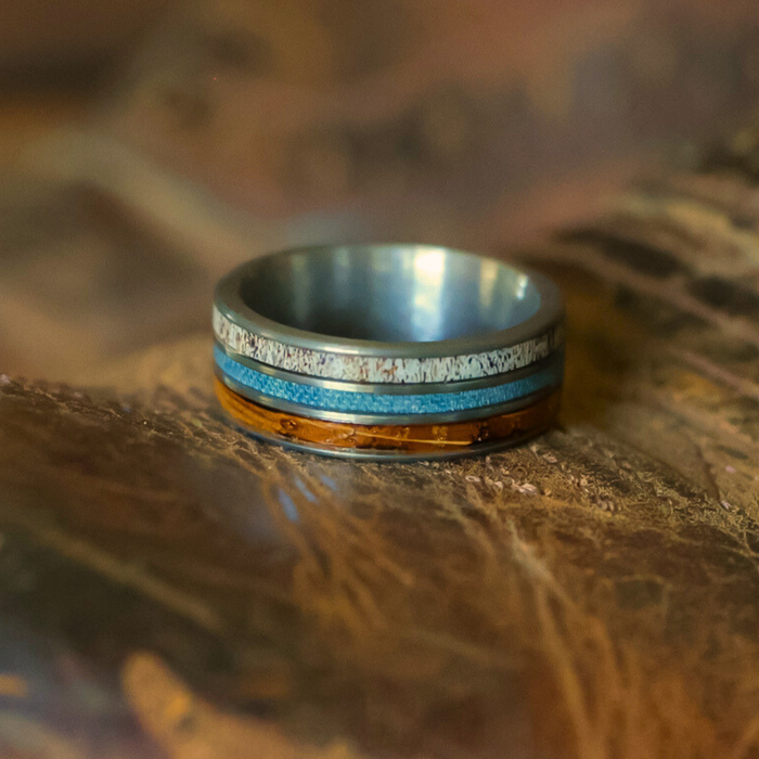 Introducing The Bodacious: a striking men's band showcasing three distinct layers—a tan speckled band on top, a slim blue stripe at the center, and a wood-toned band at the bottom. Crafted from durable tungsten carbide, this thoughtfully designed ring is perfect for those who appreciate unique antler rings. The Bodacious rests elegantly on a brown, textured surface.