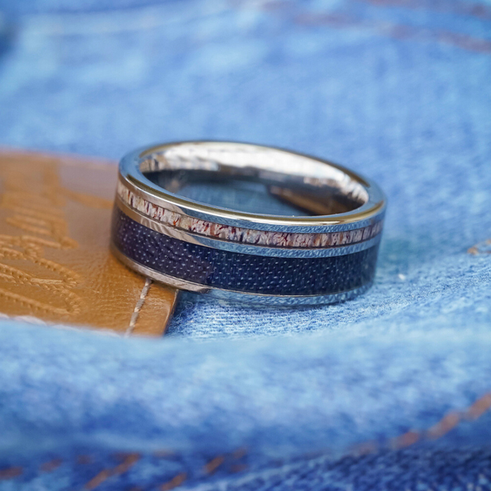 The Brazile, a silver men's band featuring a textured blue inlay, rests on tan leather atop blue denim fabric.