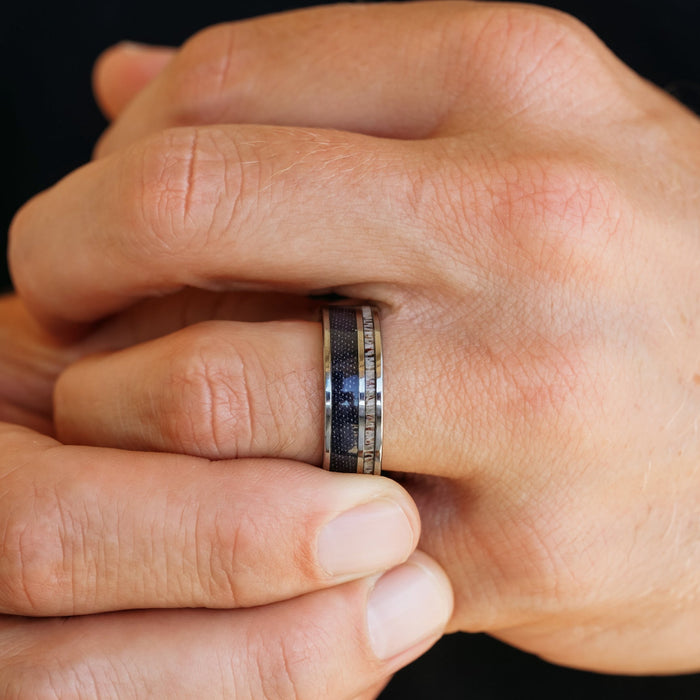Close-up of two hands clasped together, focusing on The Brazile—a men's band featuring a dark inlay and metallic edges—worn on the ring finger. The blurred background draws attention to the unique style of this tungsten carbide antler ring.