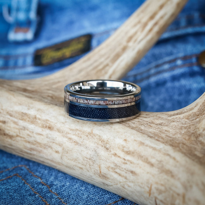 The Brazile, a silver and dark blue tungsten carbide ring, rests on a light-colored antler with blue denim fabric in the background, highlighting the rugged elegance of antler rings for men.
