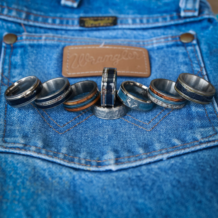Seven men’s rings, including The Brazile—a striking tungsten carbide ring—and rugged antler designs, are arranged in a row atop the back pocket of blue Wrangler jeans, with the iconic leather brand patch visible in the background.