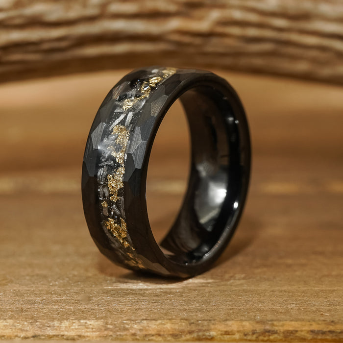 The Brenham, a black hammered ring featuring a faceted surface and a central inlay of gold flakes, rests on a brown wooden surface with a blurred wooden background.