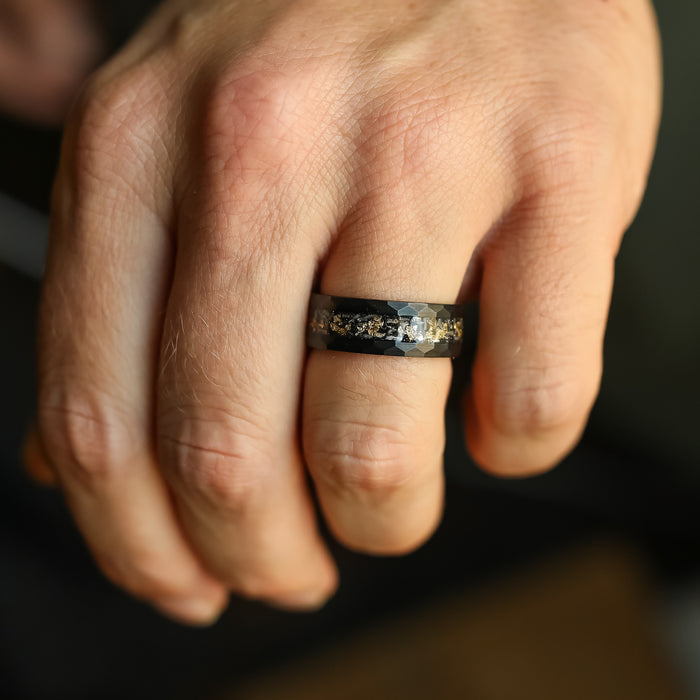 A close-up of a hand wearing The Brenham, a black hammered ring featuring a textured metallic inlay, displayed on the ring finger against a blurred background.