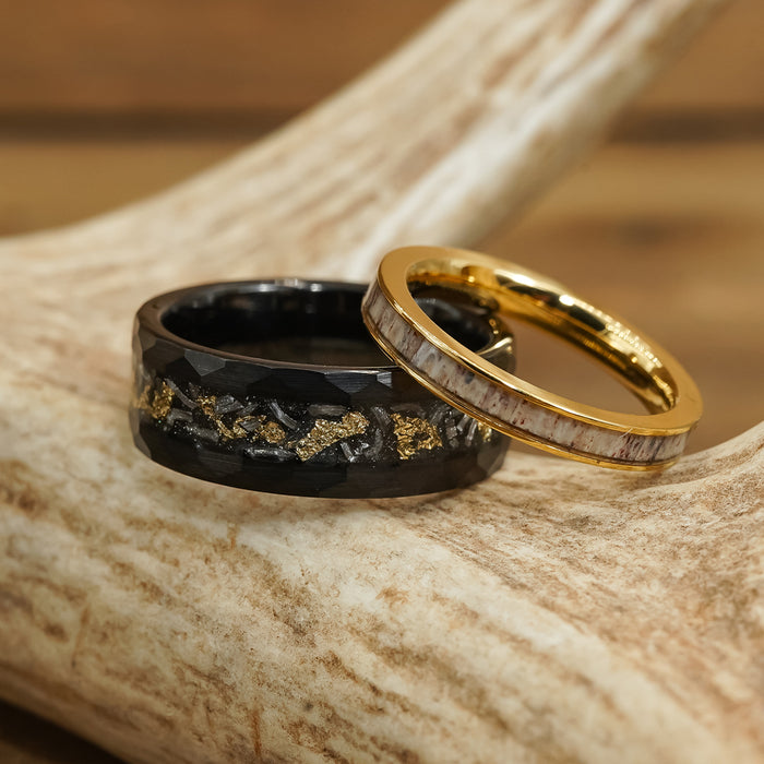 Two rings rest on a piece of antler: one is The Brenham, a black hammered ring with gold flakes and a faceted design, while the other is gold with a light, textured inlay. The background is blurred brown wood.