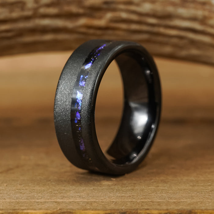 The Burro Creek, an 8mm men's black tungsten carbide ring featuring a matte finish and a striking blue-purple inlay, stands upright on a wooden surface against a blurred wooden background. This wedding band effortlessly blends modern style with timeless elegance.